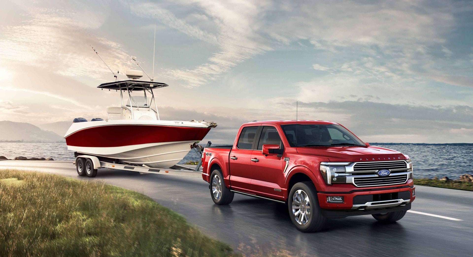 A red Ford F-150 Platinum towing a large white and red center console boat on a trailer down a road next to the water, with mountains in the background under a cloudy sky.