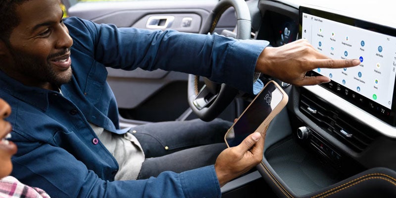 A man uses a smart car console screen by tapping icons while holding a smartphone; a passenger is barely visible.