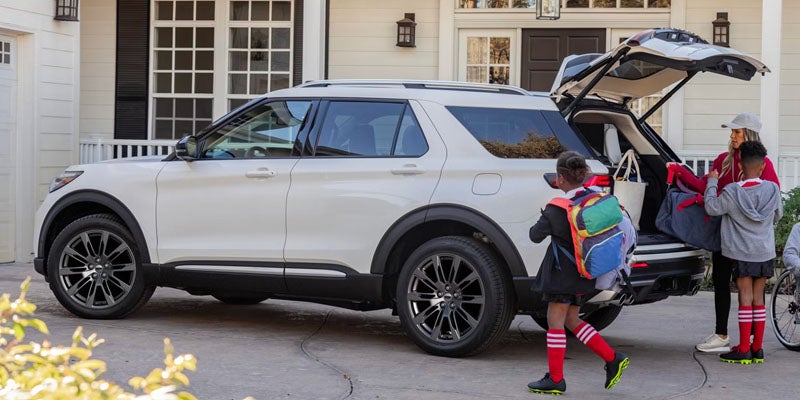 White SUV with open trunk, a woman and two children loading bags, with a house in the background.