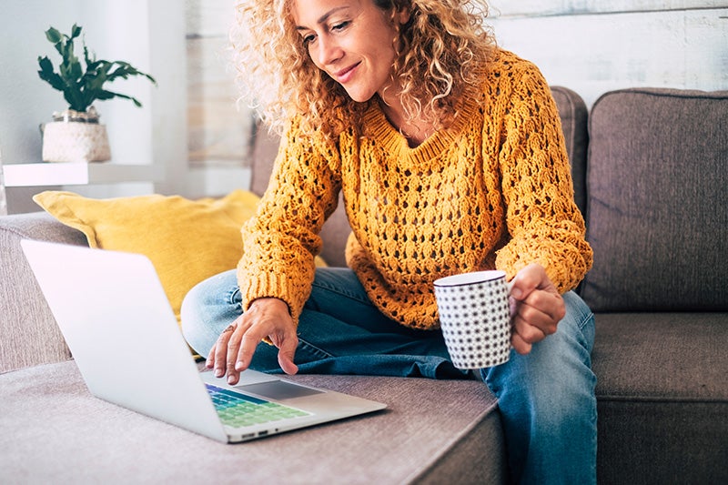 Woman with curly hair in an orange sweater and blue jeans sitting on a couch using a laptop, holding a mug.