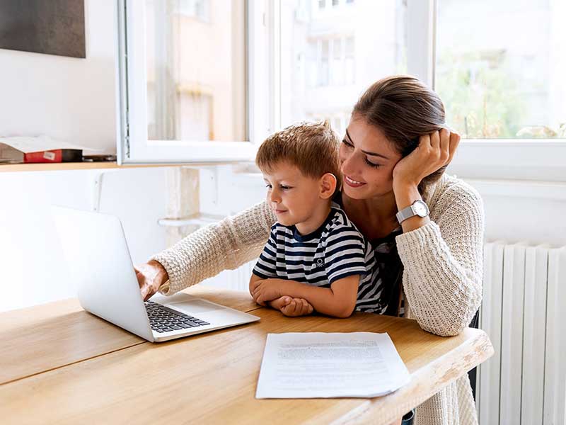 A smiling woman and a young boy sitting together at a wooden table, looking at a laptop.