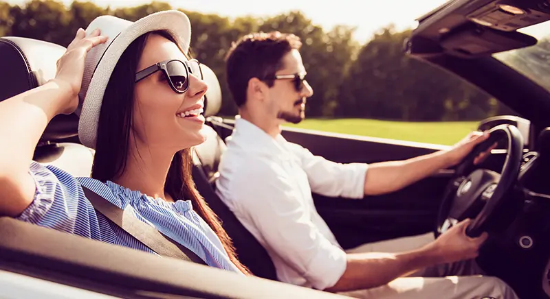 A smiling woman in a hat and sunglasses rides in a convertible with a man driving.